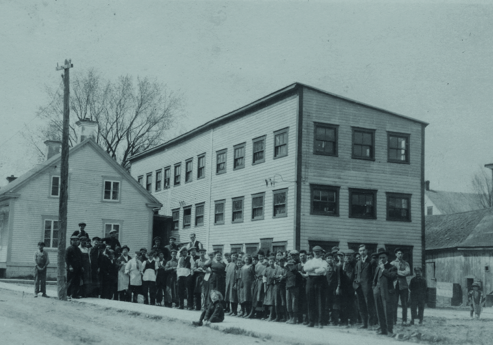 The picture shows employees assembles in front of the Charron factory. It is a three-story wooden structure, built behind a house on St-Antoine Street.