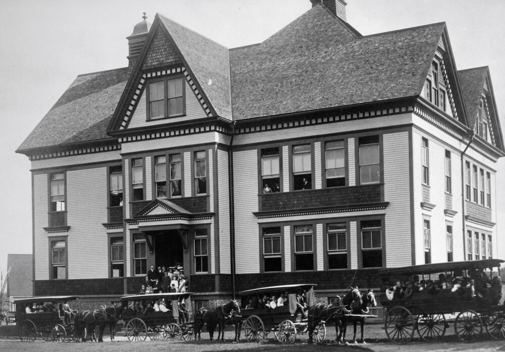 People stand on school steps. Others look out second storey windows. Students are in four school vans of varying sizes in front of the building. Each van has a driver and is led by two horses.