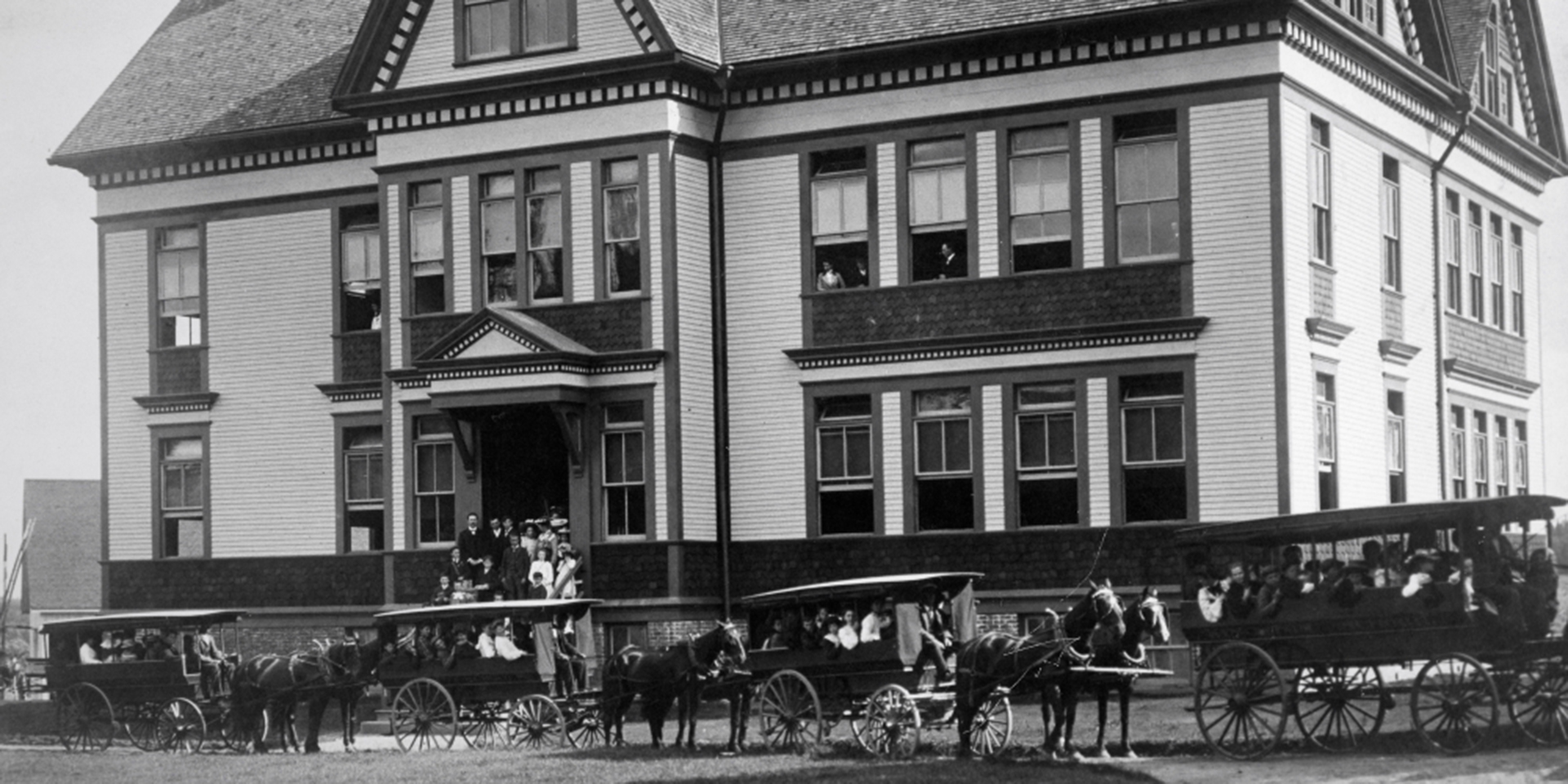 People stand on school steps. Others look out second storey windows. Students are in four school vans of varying sizes in front of the building. Each van has a driver and is led by two horses.