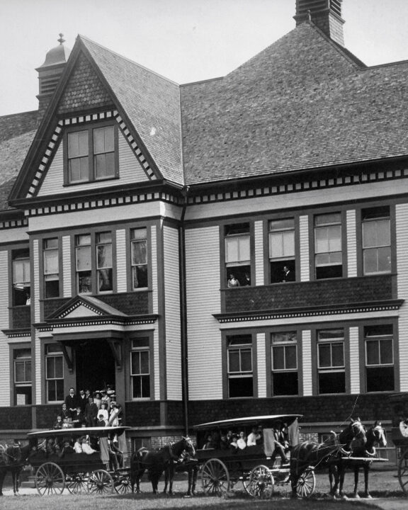 People stand on school steps. Others look out second storey windows. Students are in four school vans of varying sizes in front of the building. Each van has a driver and is led by two horses.