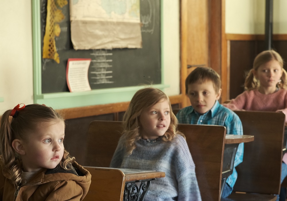 Four children sit in a row of five wooden desks in an old schoolhouse.