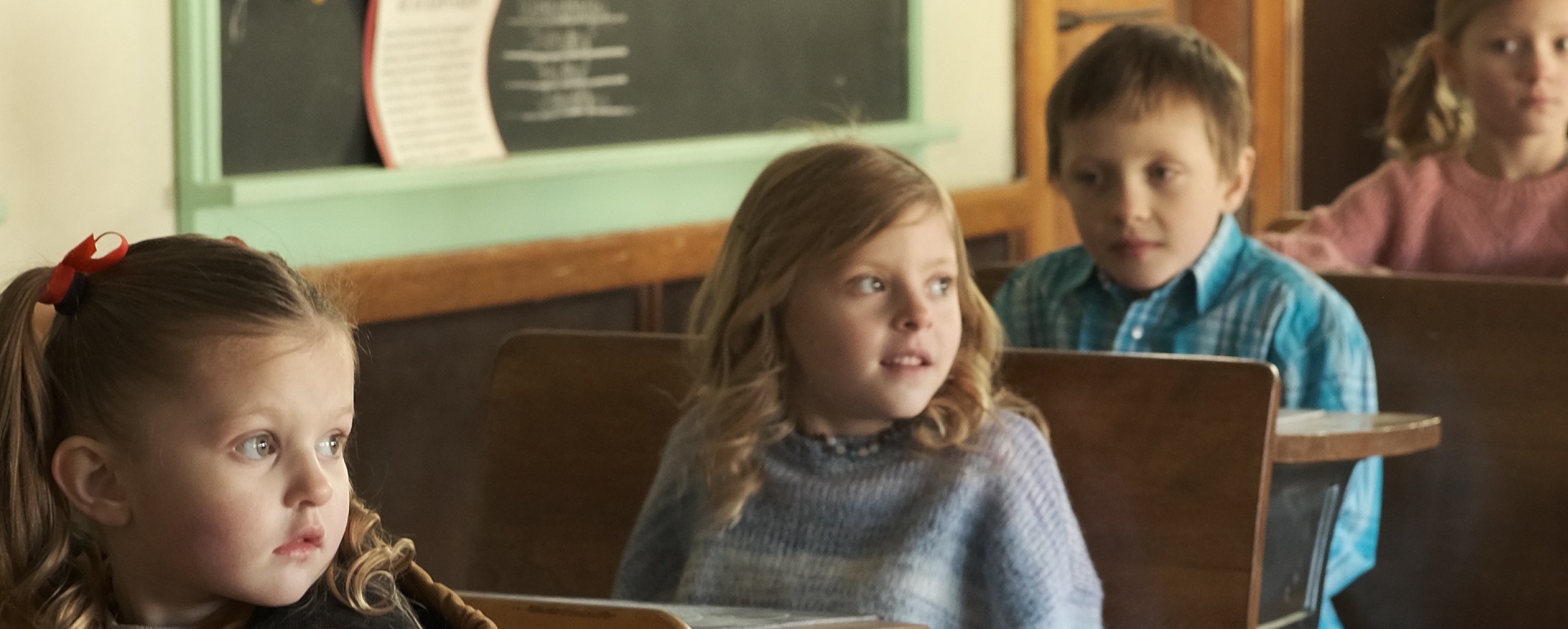 Four children sit in a row of five wooden desks in an old schoolhouse.