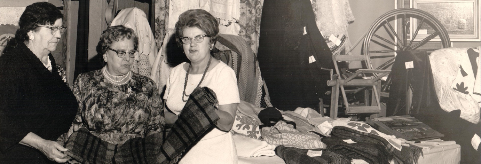 A black and white photo of three women inspecting a blanket. A quilt and some items of clothing are hanging behind them. On their right, blankets have been spread out on a table and a spinning wheel is displayed.