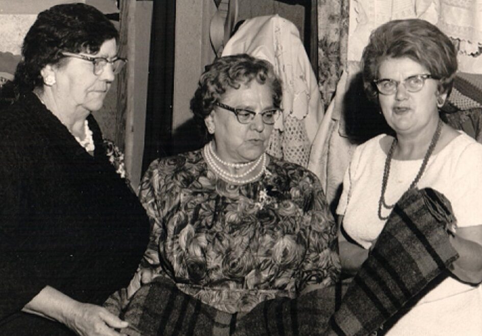 A black and white photo of three women inspecting a blanket. A quilt and some items of clothing are hanging behind them. On their right, blankets have been spread out on a table and a spinning wheel is displayed.