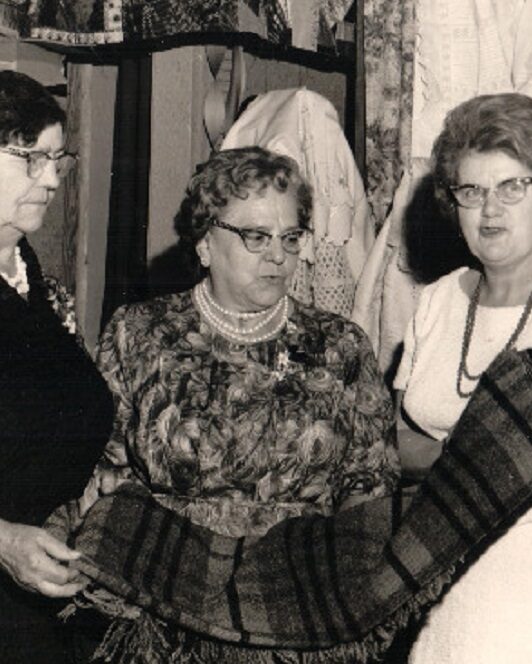 A black and white photo of three women inspecting a blanket. A quilt and some items of clothing are hanging behind them. On their right, blankets have been spread out on a table and a spinning wheel is displayed.