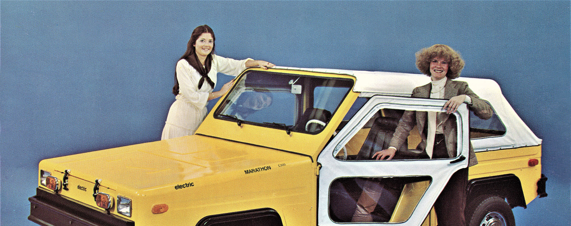 Two smiling women stand in the doors of a convertible two-seater electric truck.