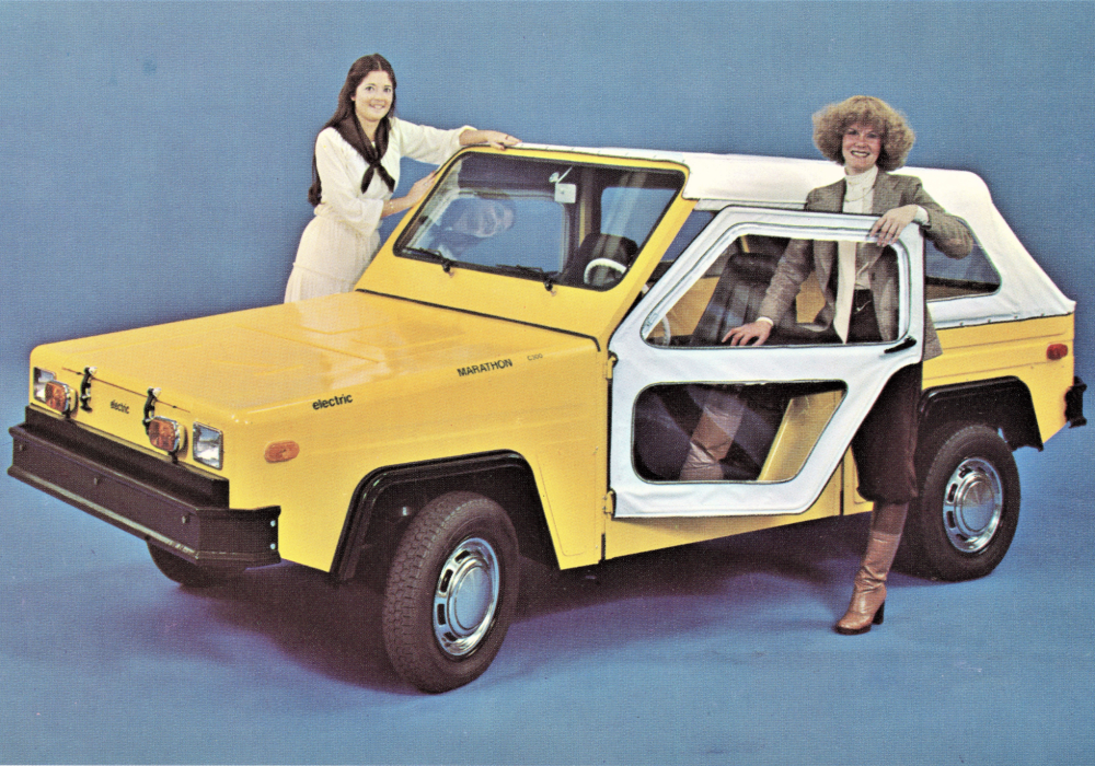 Two smiling women stand in the doors of a convertible two-seater electric truck.