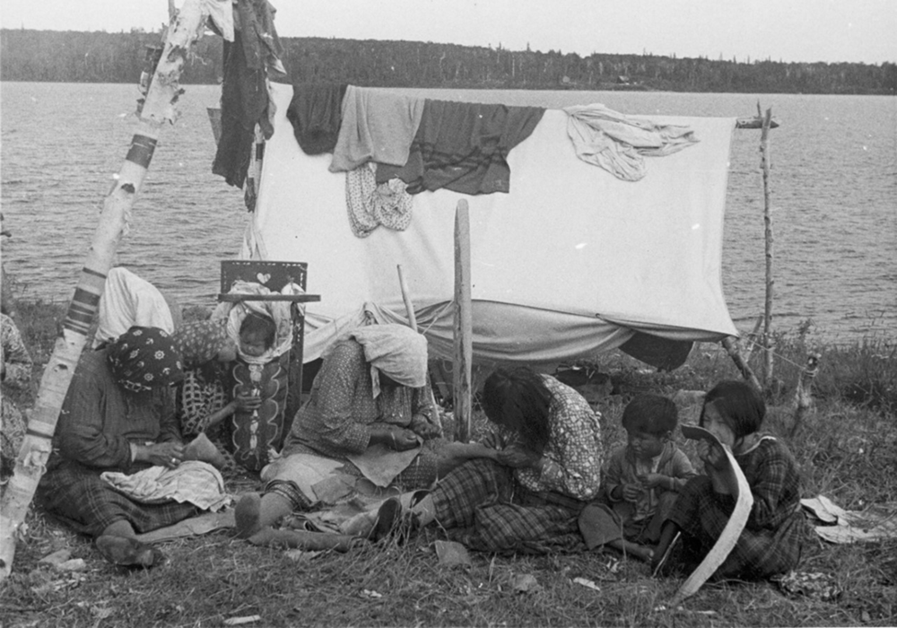 The photo is in black and white. Anicinabek women and children are sitting on the grass. Lake is in the background. They are near a small canvas tent. The women are busy sewing moccasins while the children watch or play. A baby is sitting in a tikinagan, a traditional baby carrier.