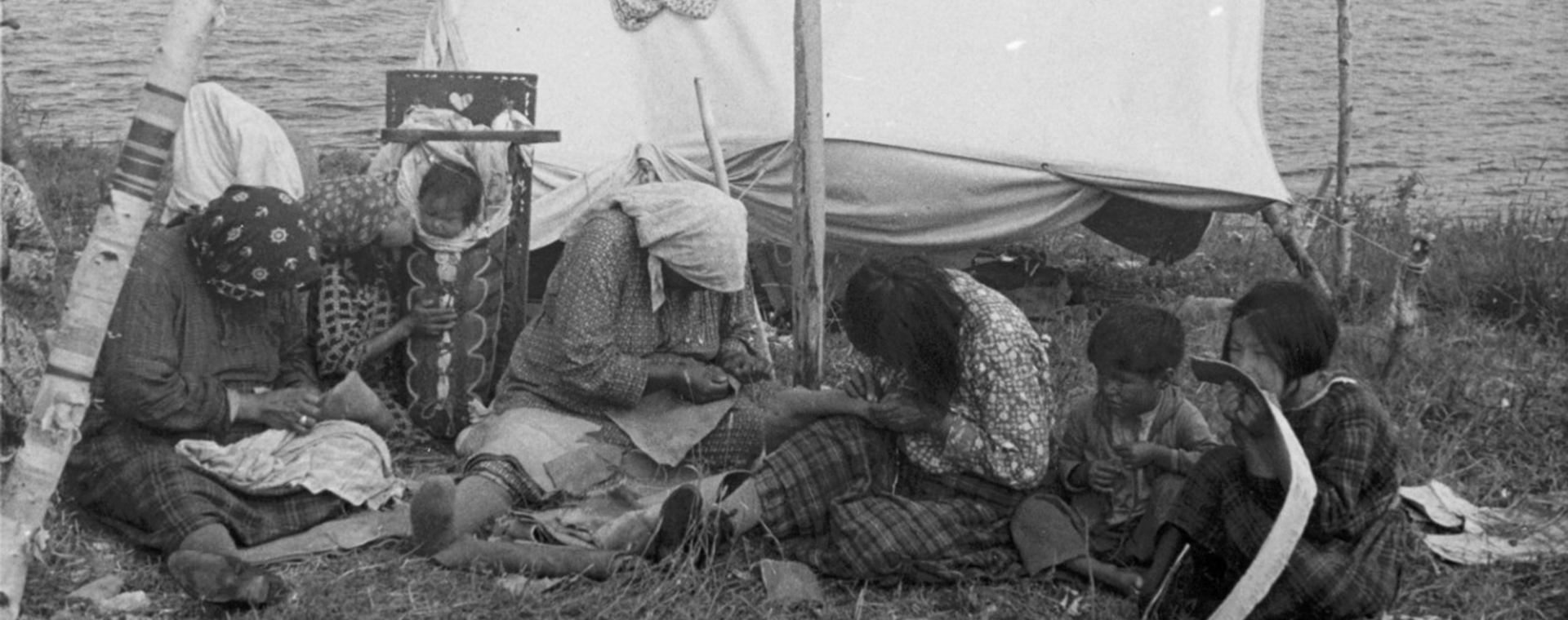 The photo is in black and white. Anicinabek women and children are sitting on the grass. Lake is in the background. They are near a small canvas tent. The women are busy sewing moccasins while the children watch or play. A baby is sitting in a tikinagan, a traditional baby carrier.