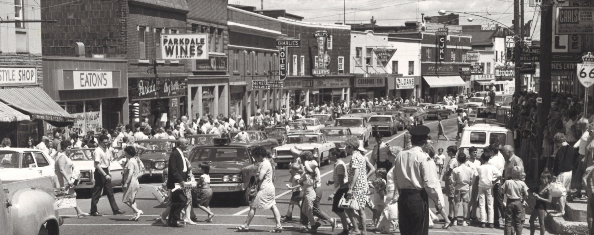 Black and white photograph of a busy street. A policeman directs traffic in the foreground while people cross the street. Cars line the street and businesses are in the background. A banner hangs over the street.