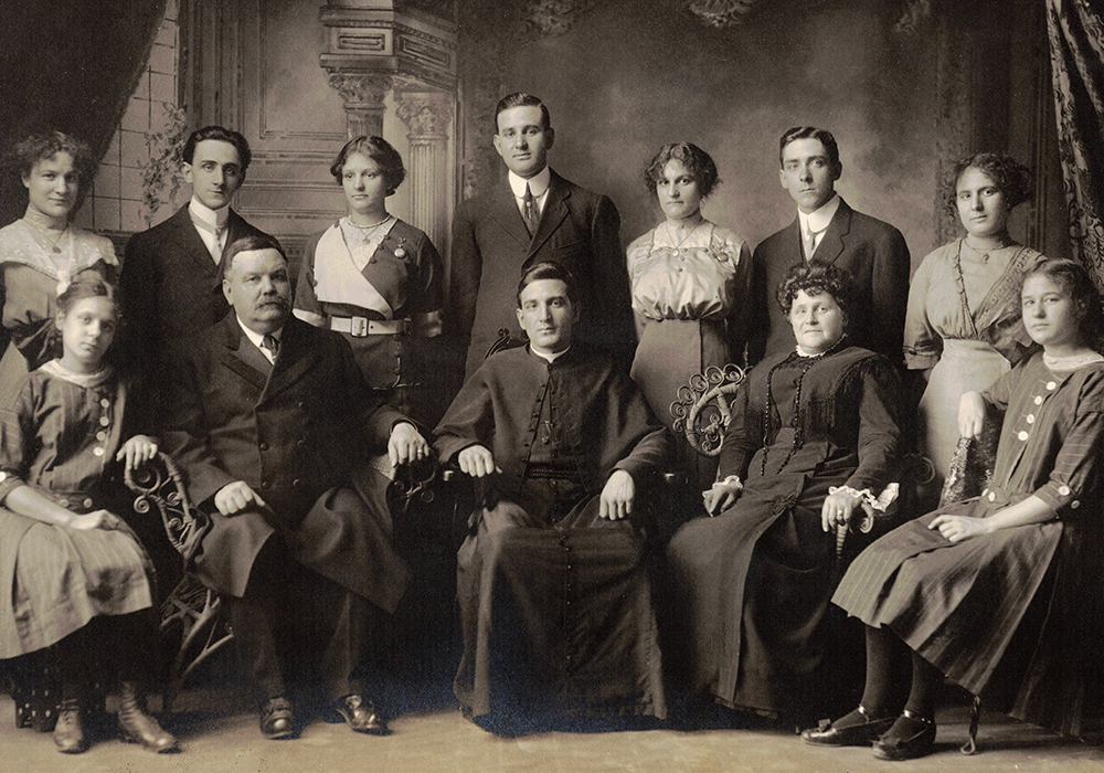 Black and white picture taken in studio. 12 people in their early 20th century clothes. In the front are seated Monsignor Alfred LePailleur, his parents and his two youngest sisters. Standing in the back are the other members of the siblings.