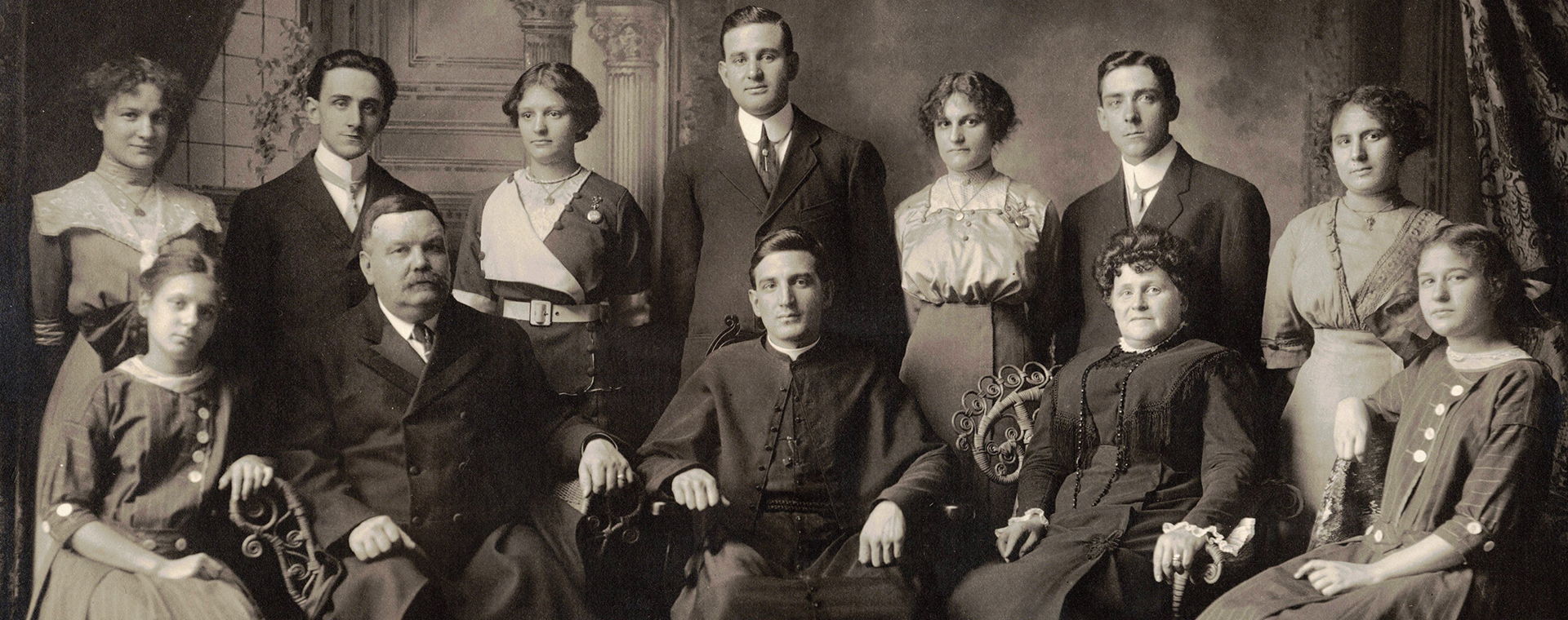 Black and white picture taken in studio. 12 people in their early 20th century clothes. In the front are seated Monsignor Alfred LePailleur, his parents and his two youngest sisters. Standing in the back are the other members of the siblings.