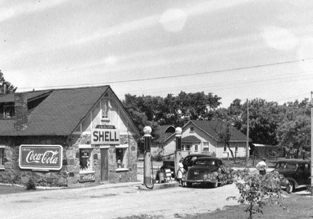 Black-and-white image of vintage cars parked in front of gas pumps with stone building in background