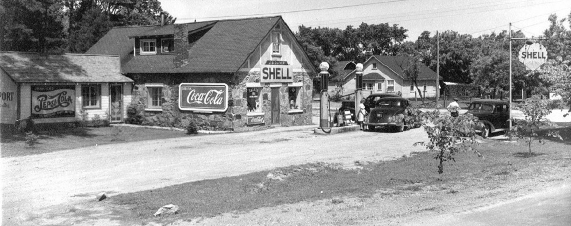 Black-and-white image of vintage cars parked in front of gas pumps with stone building in background