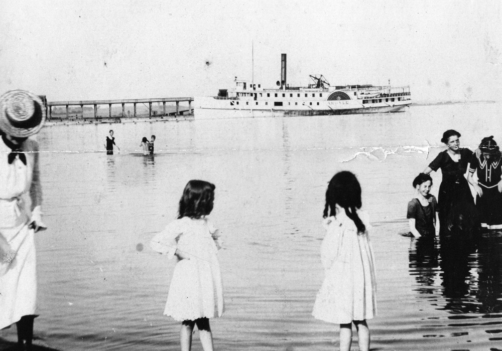 Black and white photograph of a lake with individuals standing in the water and in the background a large steamship and pier.