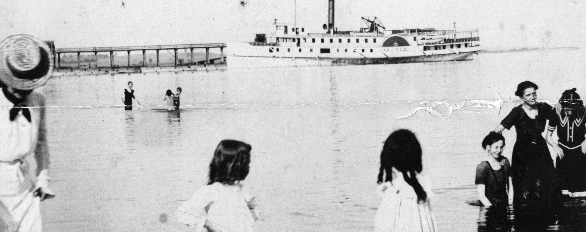 Black and white photograph of a lake with individuals standing in the water and in the background a large steamship and pier.