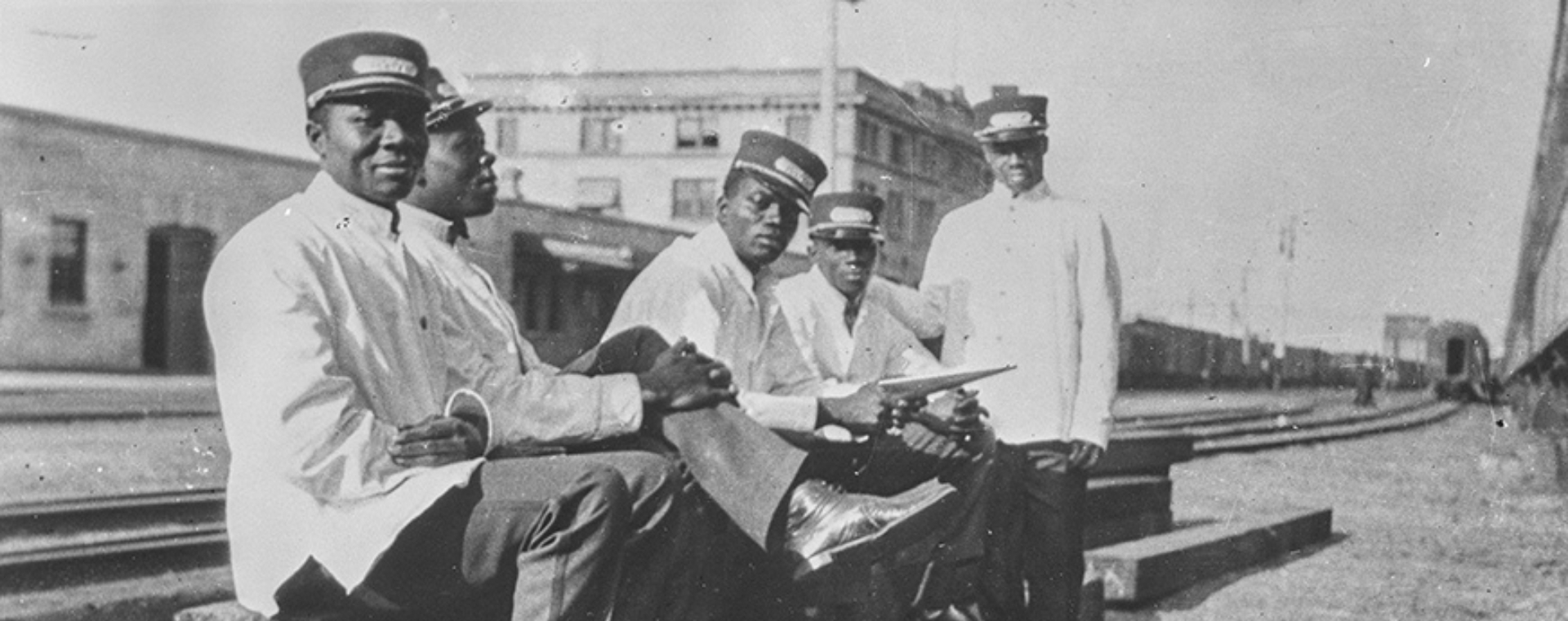 Black and white archival photograph of 5 Black men sitting on a railway track. They are dressed in white railway uniforms.