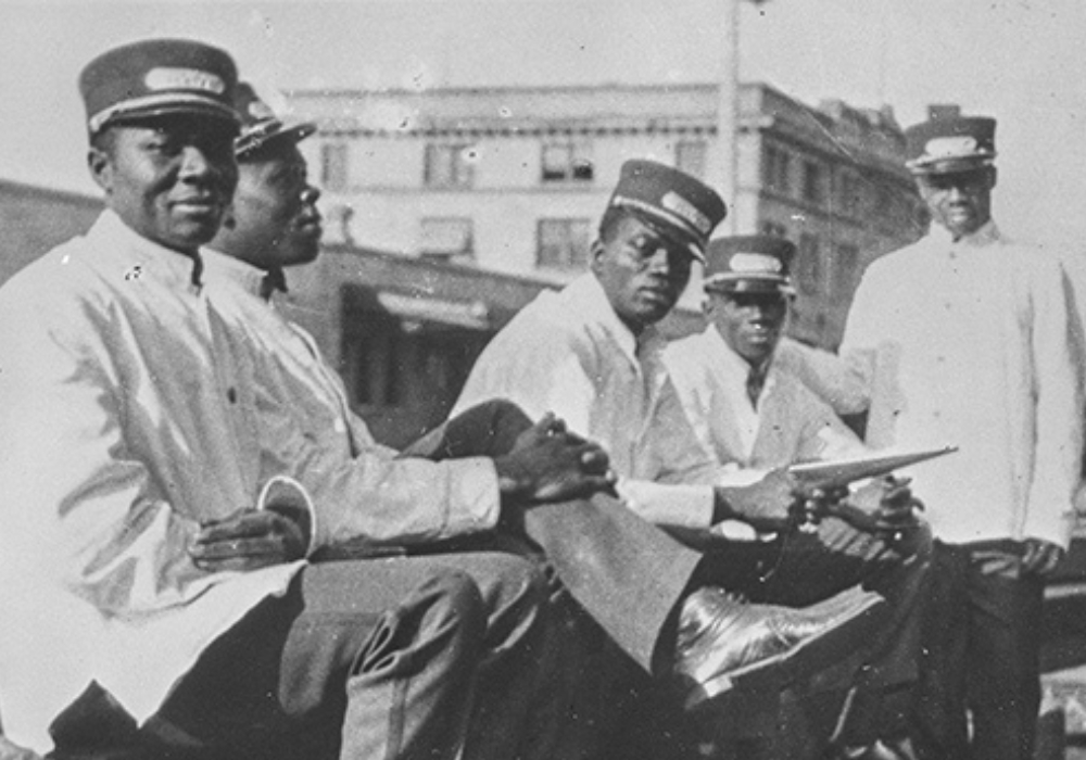 Black and white archival photograph of 5 Black men sitting on a railway track. They are dressed in white railway uniforms.