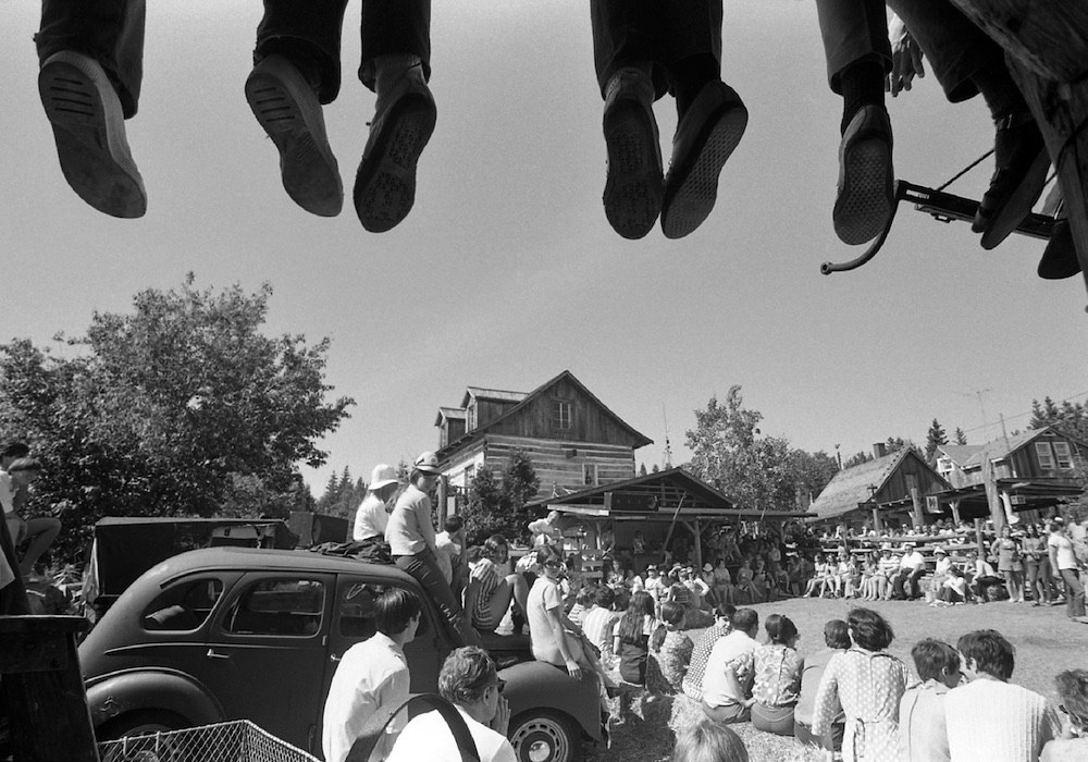 Black and white photo of about 100 spectators attending an outdoor performance at La Butte. At the top of the picture, the feet of people sitting on part of the roof of La Butte can be seen.