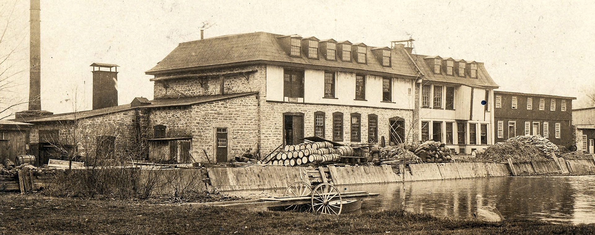 Postcard published around 1900. In the center: a paper mill, on the left: its tall smokestack overlooking the mill’s dike, cluttered with barrels and other materials. In the foreground: a roll-in dock on the water basin.