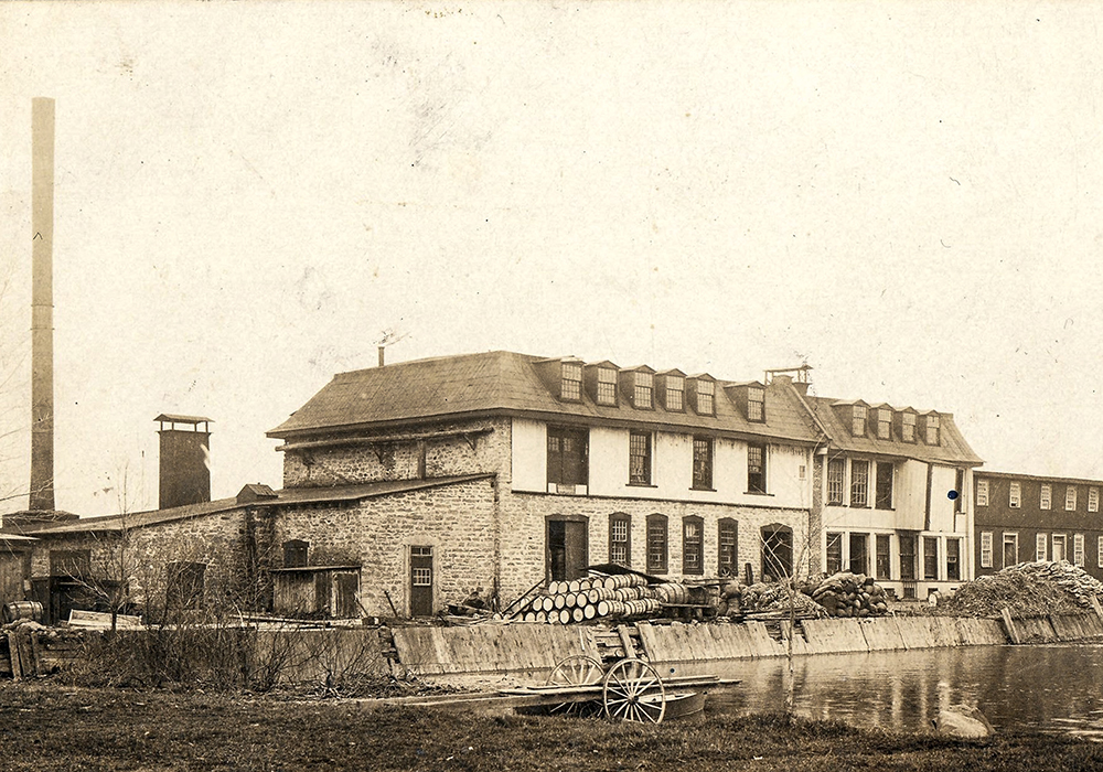 Postcard published around 1900. In the center: a paper mill, on the left: its tall smokestack overlooking the mill’s dike, cluttered with barrels and other materials. In the foreground: a roll-in dock on the water basin.