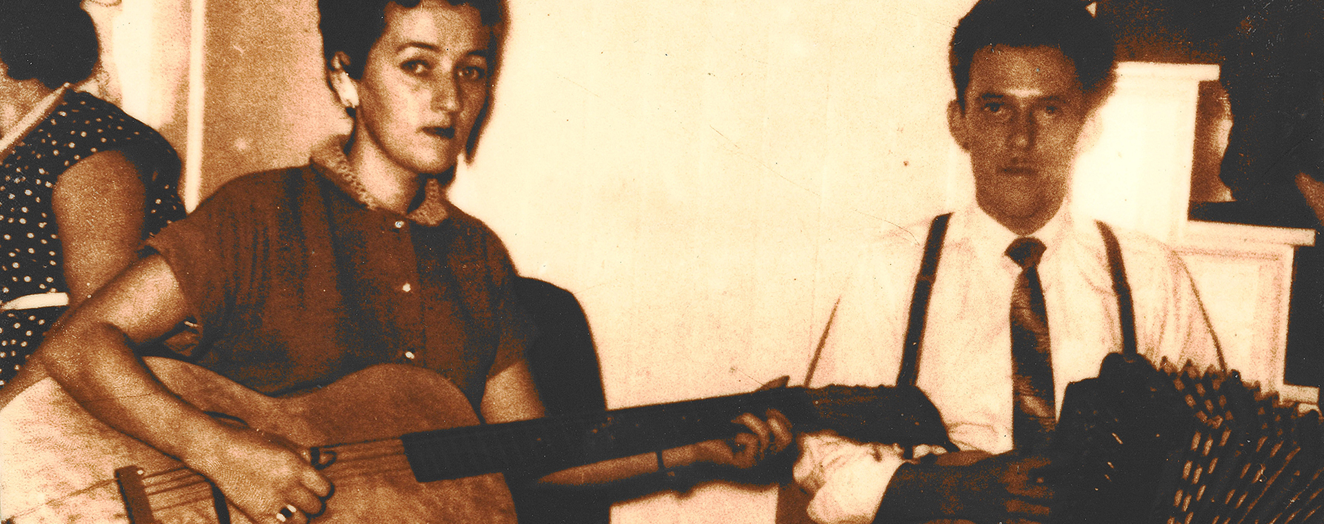 Yvette Coulombe is sitting on a chair with a guitar. Ulric Lacombe is at her right. He is sitting with a diatonic accordion on his knee