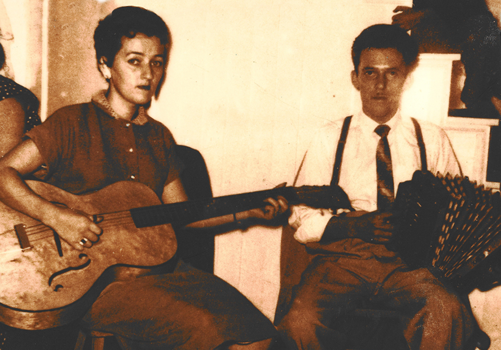 Yvette Coulombe is sitting on a chair with a guitar. Ulric Lacombe is at her right. He is sitting with a diatonic accordion on his knee