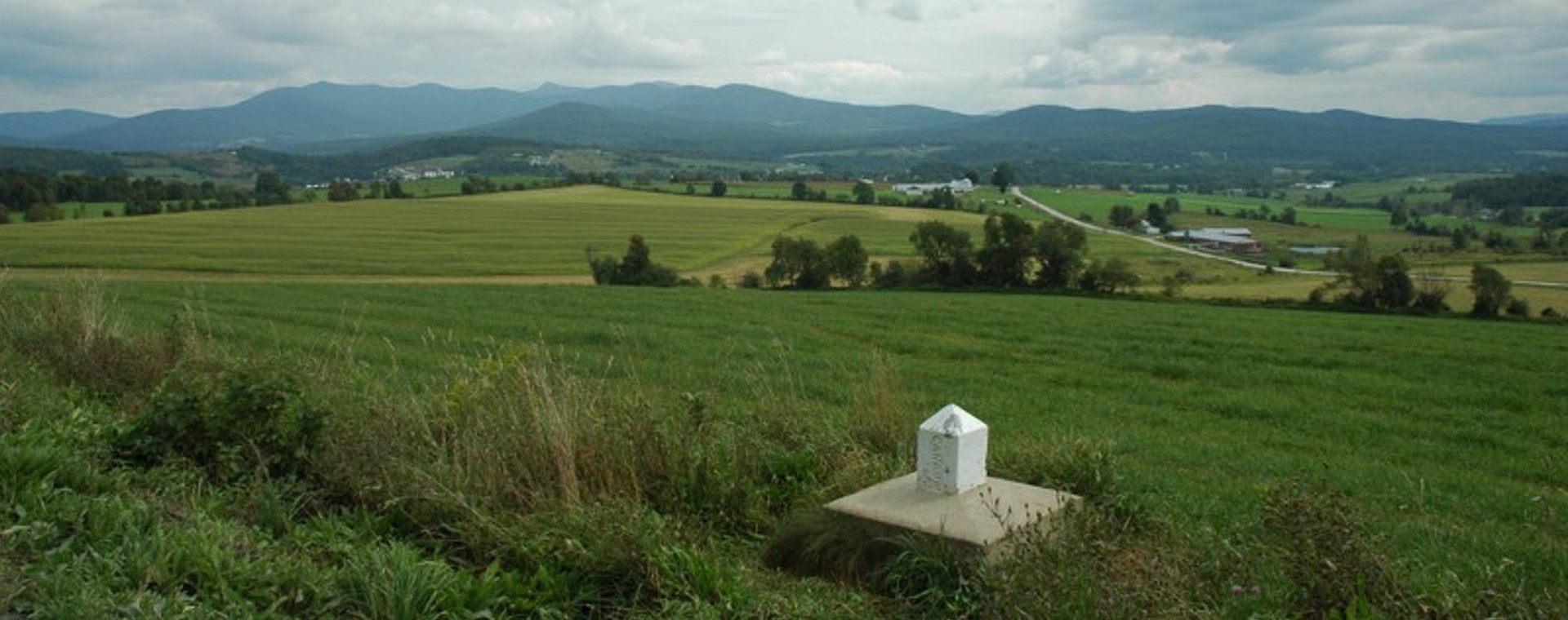In the middle of a leafy field at the foot of the mountains, a small white marker indicates the Canada-U.S. border.