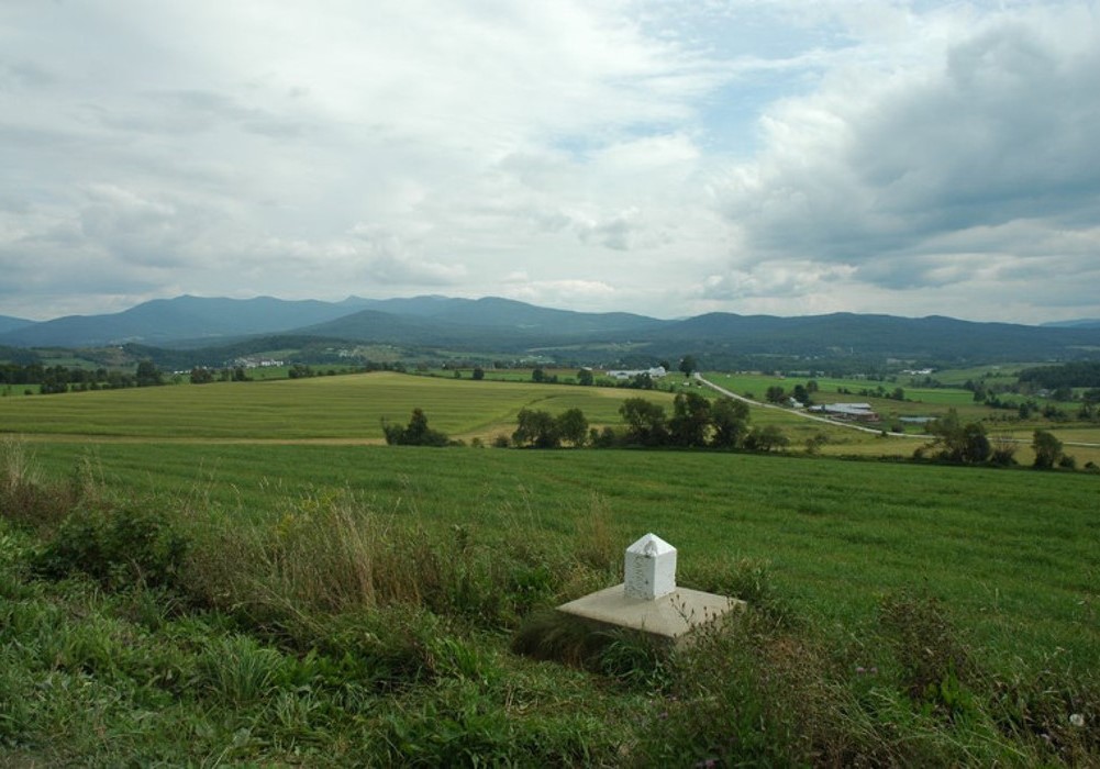 In the middle of a leafy field at the foot of the mountains, a small white marker indicates the Canada-U.S. border.