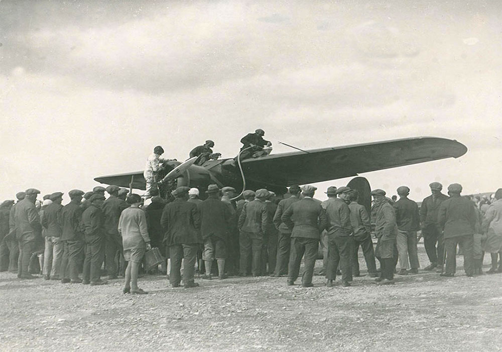 A black and white photo of three individuals on top of the wings of a plane, refueling. A large crowd stood around the aircraft, backs to camera.