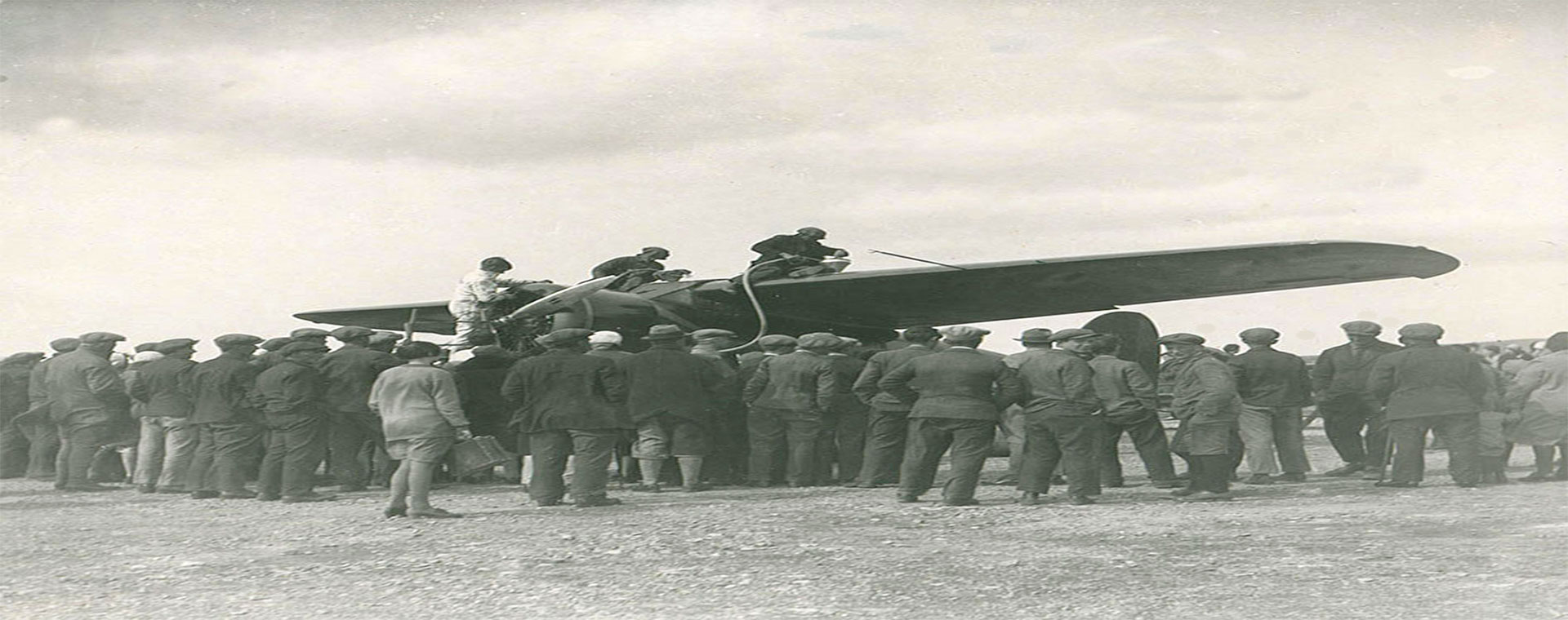 A black and white photo of three individuals on top of the wings of a plane, refueling. A large crowd stood around the aircraft, backs to camera.