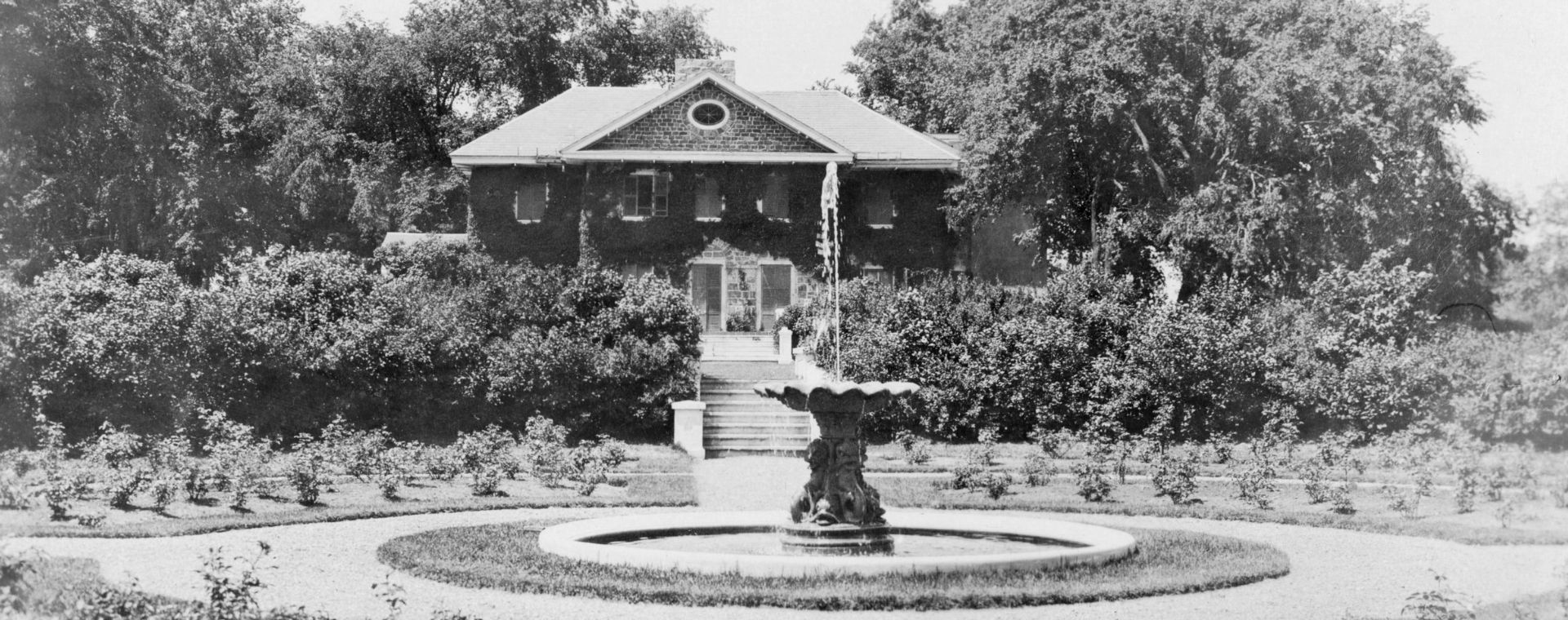 Black and white photograph of a circular garden of shrubs surrounding a fountain in front of a large two-story stone house covered in climbing plants and with a staircase leading to the garden.