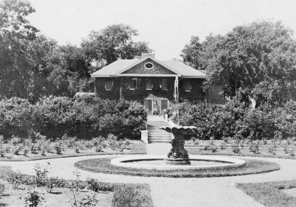 Black and white photograph of a circular garden of shrubs surrounding a fountain in front of a large two-story stone house covered in climbing plants and with a staircase leading to the garden.
