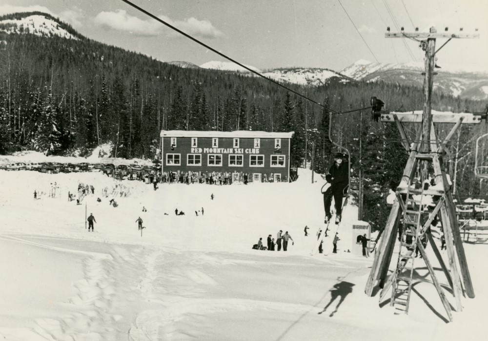 A black-and-white photo of a skier riding up a chair lift. In the distance is the Red Mountain Ski Club Lodge.