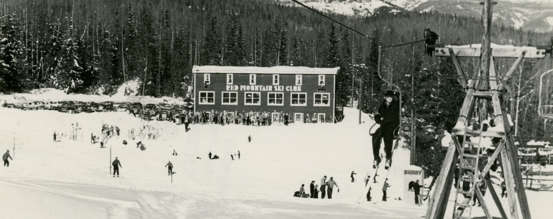 A black-and-white photo of a skier riding up a chair lift. In the distance is the Red Mountain Ski Club Lodge.
