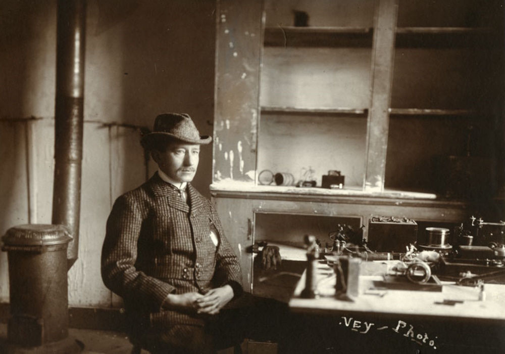 A man in a checked jacket and hat sits at a table topped by wireless equipment. Behind him are bare cupboards with no doors, and a metal stove and stovepipe.
