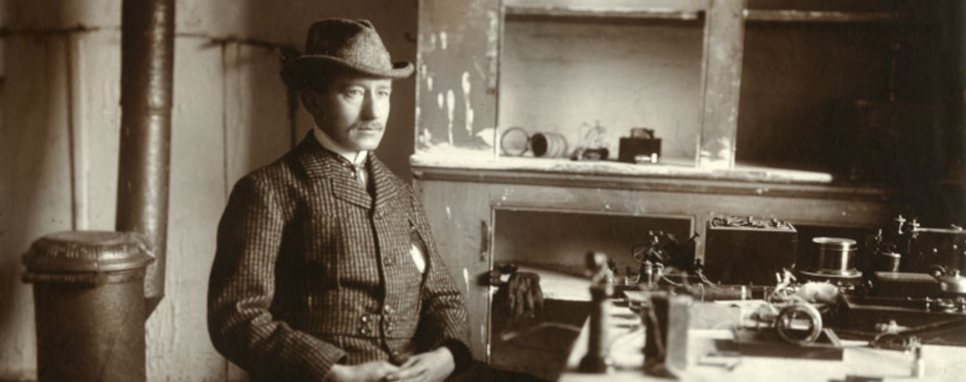 A man in a checked jacket and hat sits at a table topped by wireless equipment. Behind him are bare cupboards with no doors, and a metal stove and stovepipe.