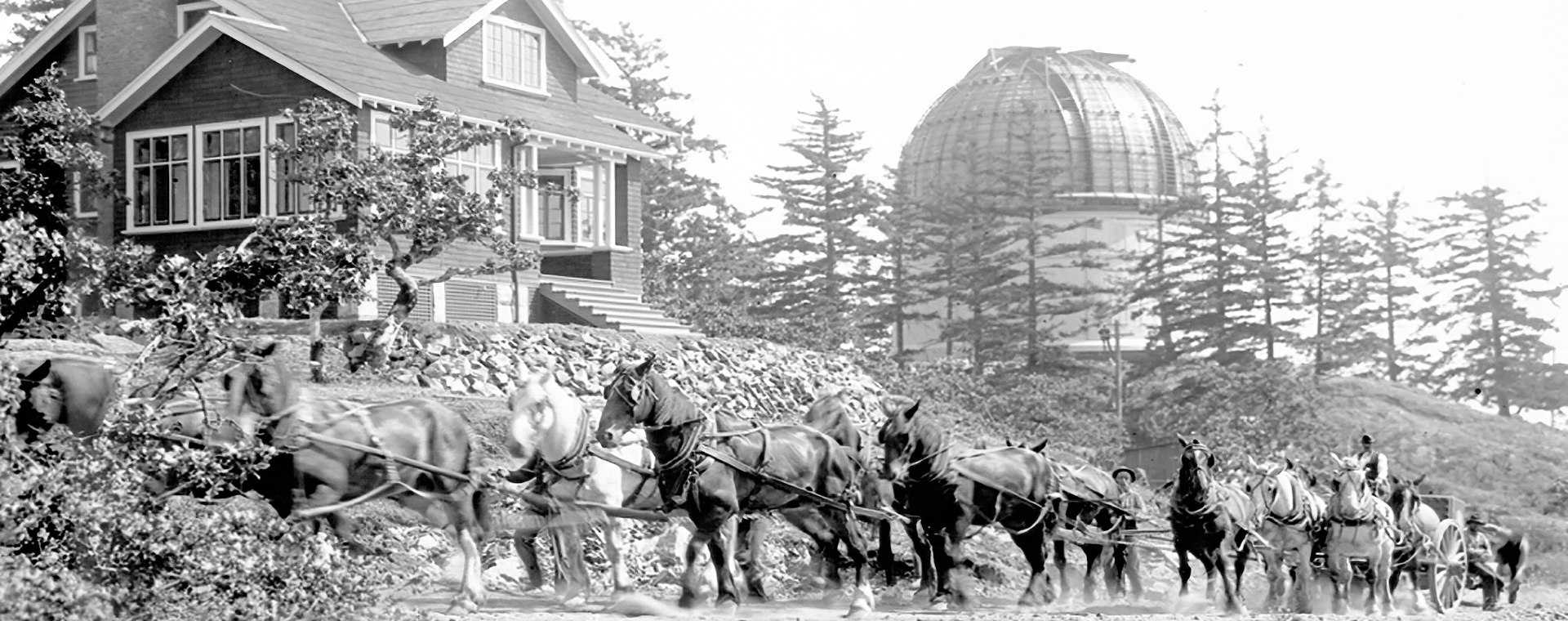 A black and white photograph showing part of a 1.8 m telescope being hauled by horse-drawn wagon, with telescope dome and the Director’s residence in background