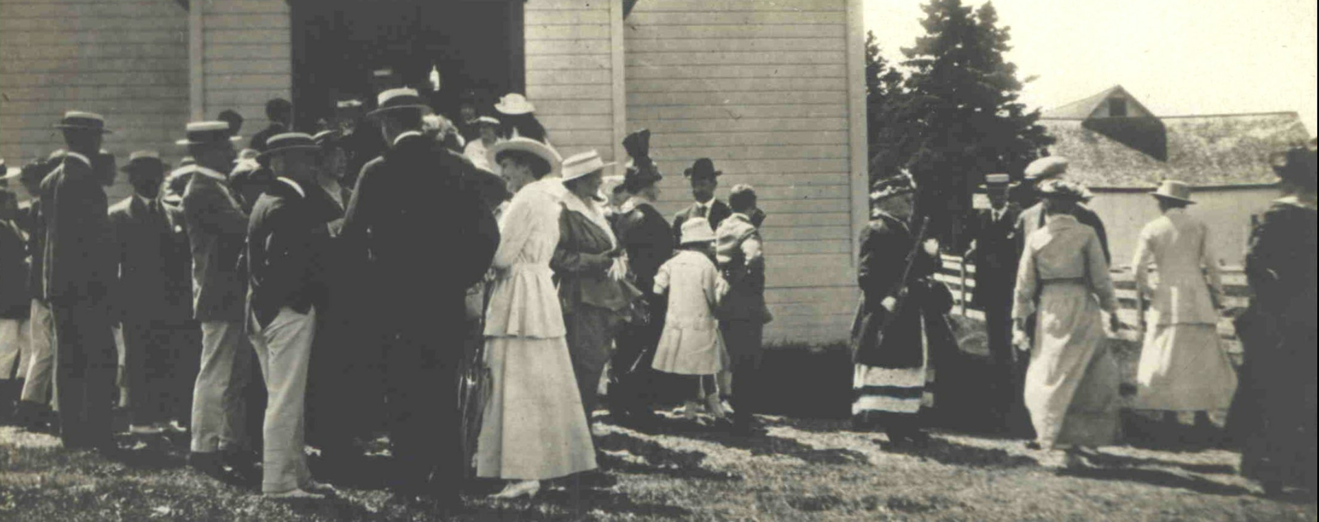 Photographie noir et blanc d'hommes et de femmes attroupés devant un église de bois à la fin de la messe. Les hommes sont, en majorité, vêtu d'un habit au veston foncé, d'un pentalon blanc et d'un chapeau tressé plat décoré d'un ruban. Les femmes portent une longue robe blanche et un chapeau plat tressé. En arrière plan, on aperçoit derrière un massif d'épinettes matures, un bâtiment de ferme.