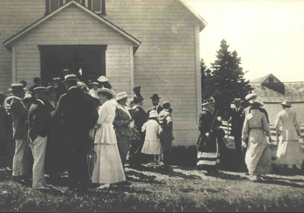 Black and white photograph of men and women gathered in front of a wooden church at the end of mass. The men are, for the most part, dressed in a dark jacket, a white pentalon and a flat braided hat decorated with a ribbon. The women wear a long white dress and a flat braided hat. In the background, a farm building can be seen behind a clump of mature spruce trees.