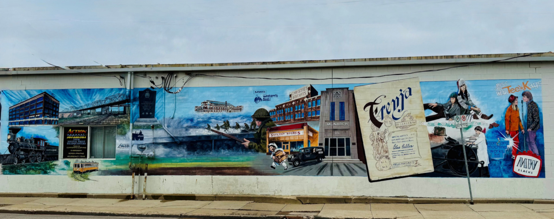 A colourful mural painted on the side of a building depicts scenes from Icelandic-Canadian history. On the left is a red brick factory marked Gimli Fish, beside a river and small figure in a white cap. At the centre stands the Icelandic newspaper building Lögberg-Heimskringla with a vintage car in front. To the right, a large image of the literary magazine Freyja shows its decorative title page, while a motorcyclist and man in old-fashioned clothing appear at the far right.