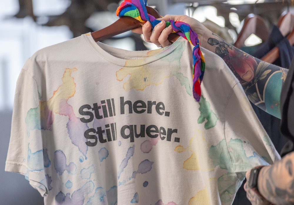 A tattooed arm is holding up an off-white t-shirt on a wooden hanger. The shirt has a multicoloured pattern of random, amorphous blobs resembling ink or watercolour. Black text across the front of the shirt reads 
