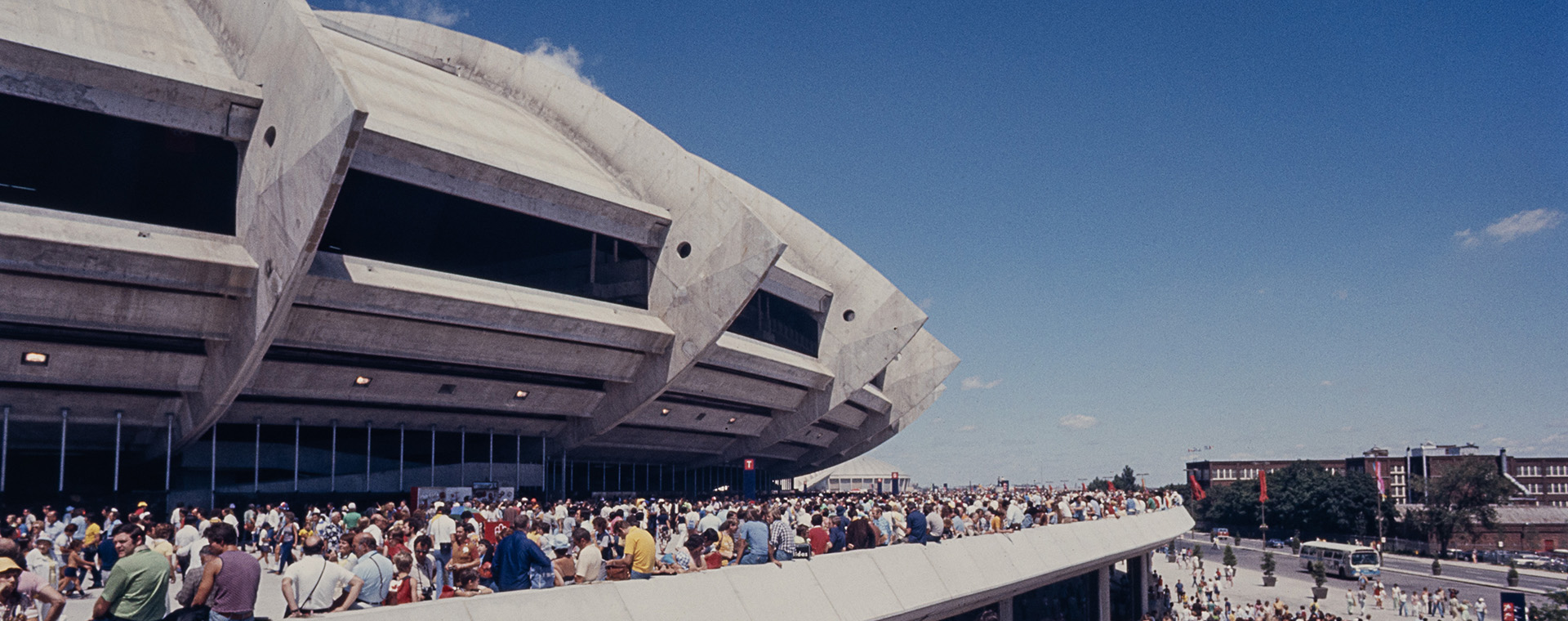 De nombreuses personnes vêtues d’habits d’été colorés se dirigent vers le Stade olympique, en empruntant une large rampe de béton. D’autres sont assises sur le rebord de l’esplanade bondée entourant le bâtiment.