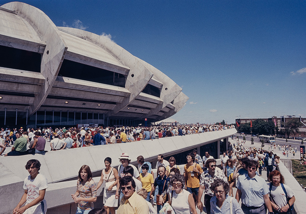 Numerous people dressed in colourful summer clothing head towards the Olympic Stadium along a wide concrete ramp. Others sit along the edge of the crowded esplanade surrounding the building.
