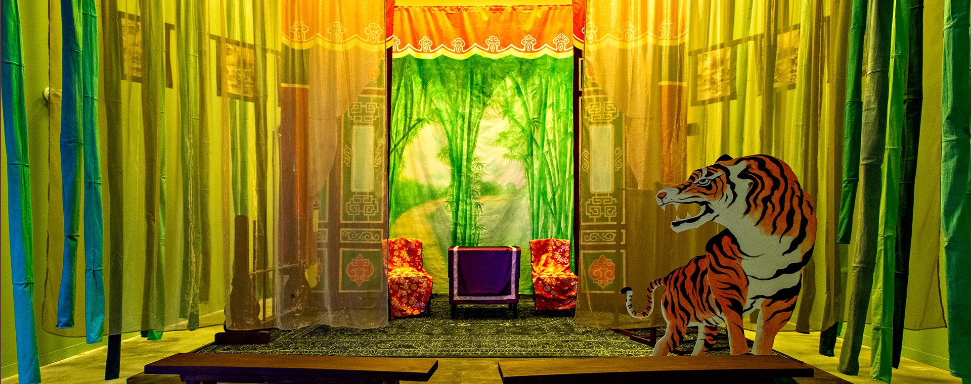View of Scent of Thunderbolts from directly in front of the stage. Wooden benches are arranged for the audience, fabric strips resembling bamboo hang on both sides, and a painted tiger cutout appears at stage right with chairs and drapes in the background.