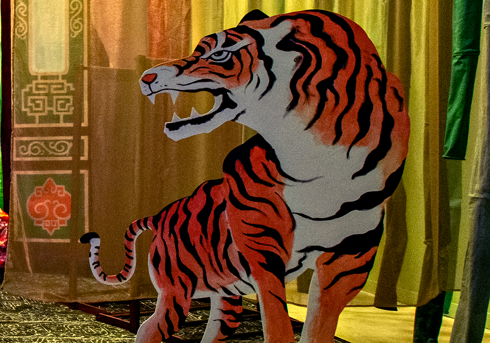 View of Scent of Thunderbolts from directly in front of the stage. Wooden benches are arranged for the audience, fabric strips resembling bamboo hang on both sides, and a painted tiger cutout appears at stage right with chairs and drapes in the background.